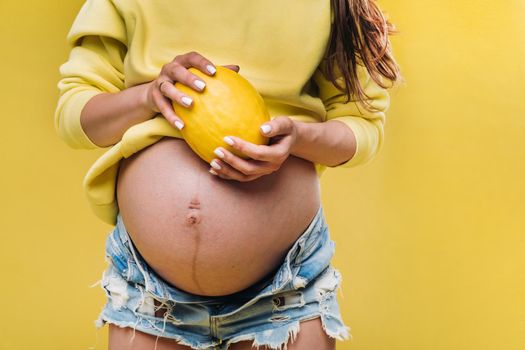 a pregnant girl with a yellow jacket stands with a melon in her hands on a yellow isolated background.