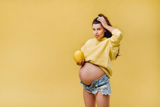 a pregnant girl with a yellow jacket stands with a melon in her hands on a yellow isolated background.