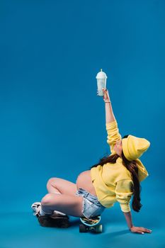 a pregnant girl in yellow clothes with a glass of juice sits on a skateboard on a blue background.