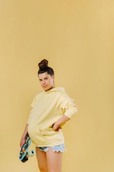 Pregnant girl in a yellow jacket with a skateboard in her hands on a yellow background.