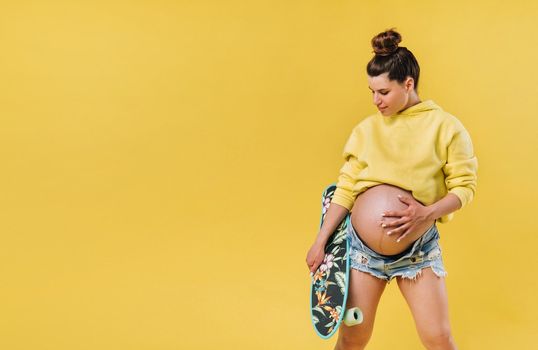 Pregnant girl in a yellow jacket with a skateboard in her hands on a yellow background.