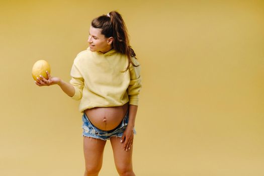 a pregnant girl with a yellow jacket stands with a melon in her hands on a yellow isolated background.