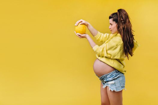 a pregnant girl with a yellow jacket stands with a melon in her hands on a yellow isolated background.