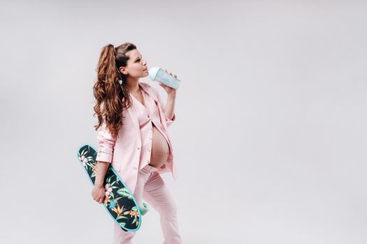 A pregnant girl in a pink suit with a skateboard in her hands drinks juice on a gray background.