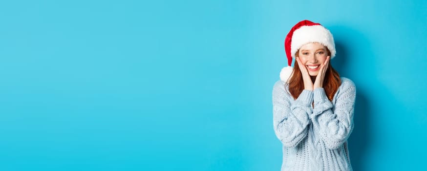 Winter holidays and Christmas Eve concept. Cheerful redhead girl in santa hat, celebrating New Year, blushing and smiling happy, standing over blue background.