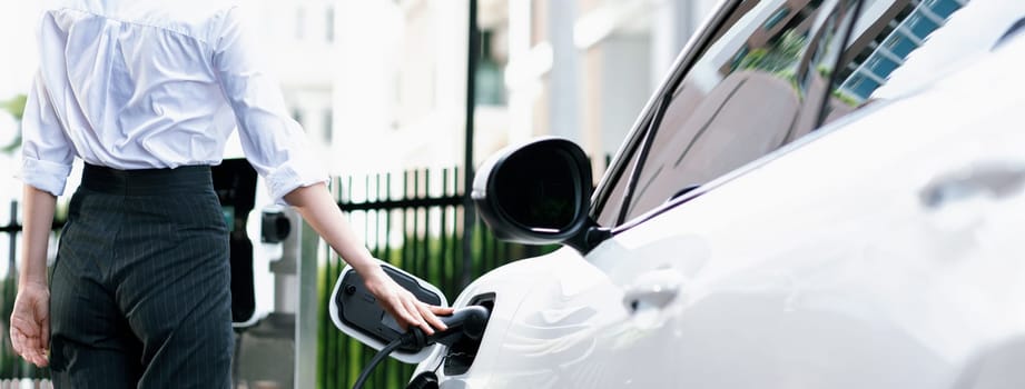 Closeup progressive suit-clad businesswoman with her electric vehicle recharge her car on public charging station in modern city with power cable plug and renewable energy-powered electric vehicle.