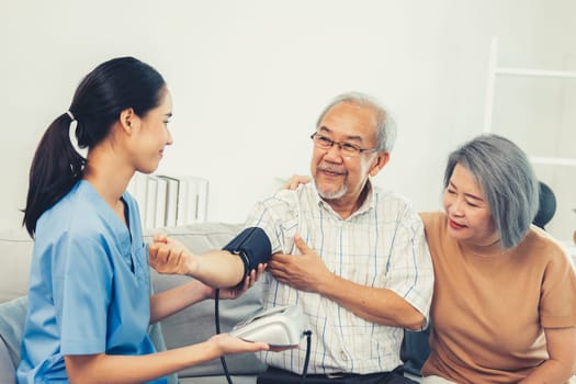 An elderly man having a blood pressure check by his personal caregiver with his wife sitting next to him in their home.