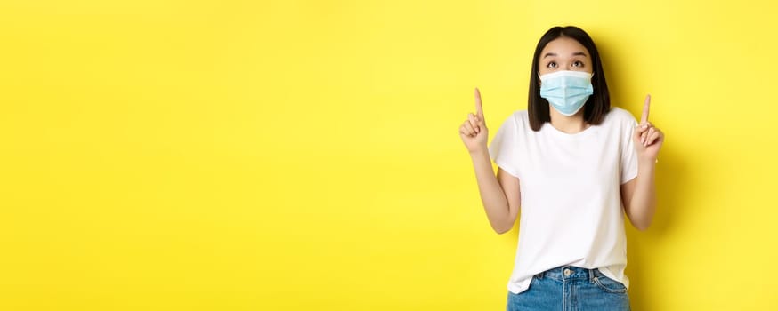 Covid-19, pandemic and social distancing concept. Young asian woman in white t-shirt and medical mask from coronavirus, looking and pointing fingers up, showing special offer.