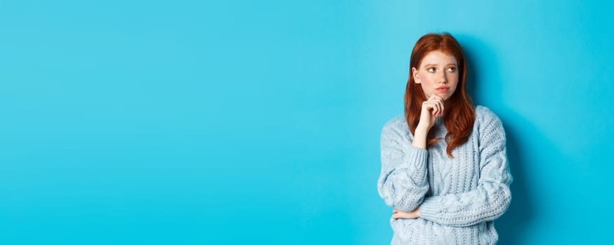 Thoughtful and upset redhead girl looking right, pondering solution, standing in sweater against blue background.