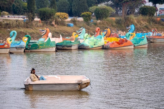 Chandigarh, India - circa 2023 : Man rowing an old damaged pedal boat on landmark sukhna lake with other boats parked in the distance showing the lives of the people running the attraction at this tourist resort