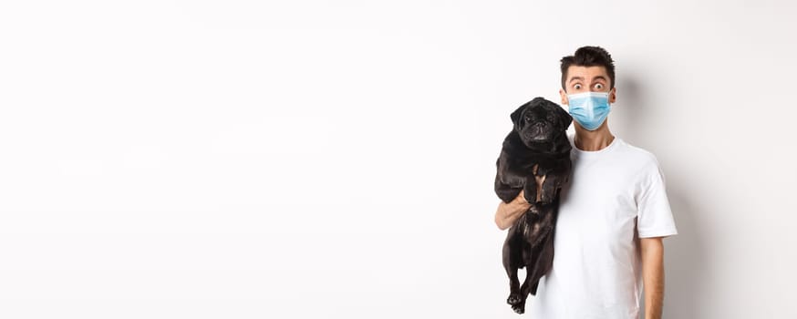 Covid-19, animals and quarantine concept. Funny young man in medical mask holding cute black pug dog, standing over white background.