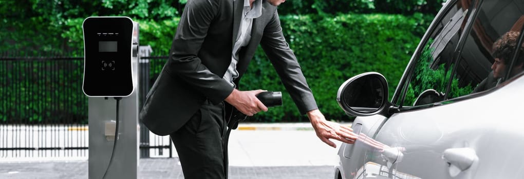 Progressive businessman with electric car recharging at public charging station at modern city residential area. Eco friendly rechargeable car powered by alternative clean energy.