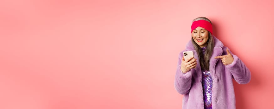 Online shopping and fashion concept. Cool asian senior woman checking out promotion on mobile phone, pointing finger at smartphone screen and smiling pleased, pink background.