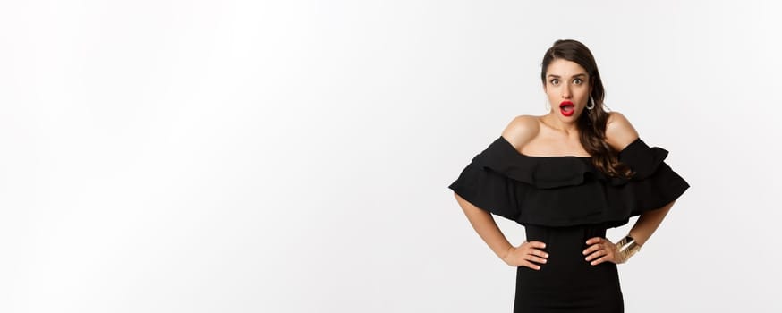 Beauty and fashion concept. Elegant woman in black dress staring at camera surprised, express complete disbelief, standing amazed over white background.