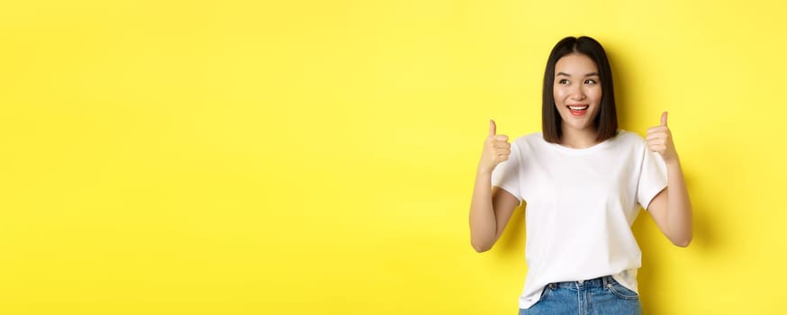 Pretty young asian woman in white t-shirt, showing thumbs up and smiling, praise good offer, recommend product, standing satisfied over yellow background.