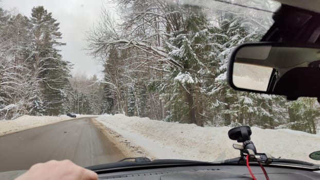 Hand of woman on the steering wheel in a car and forest and snow on the side of the road on a winter day. Woman driving car in winter travel in landscape with snow covered woods