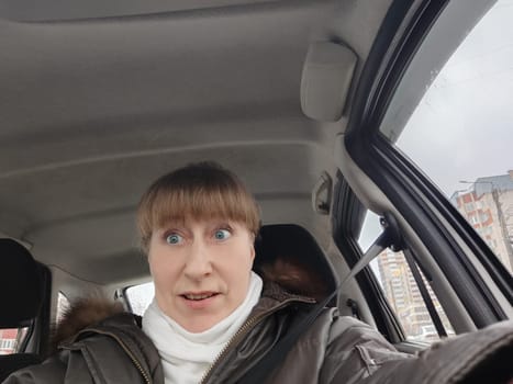 Portrait of adult cute funny blonde woman in warm clothes taking a selfie inside the car and driving it. The concept of pleasant trip in spring, autumn, winter