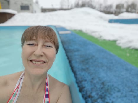 Adult mature woman takes selfie in a pool with warm hot termal mineral water in winter and white snow around. Wellness center and the concept of health care. Travel, recreation, medicine and rest