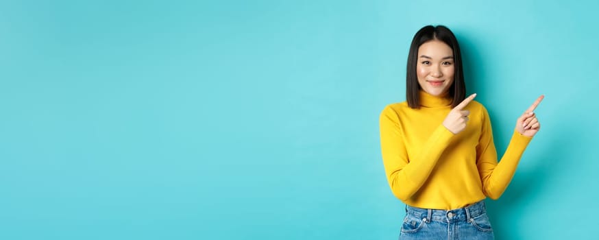 Shopping concept. Attractive young asian woman with perfect skin, wearing trendy yellow sweater and jeans, pointing fingers right and smiling, showing advertisement.