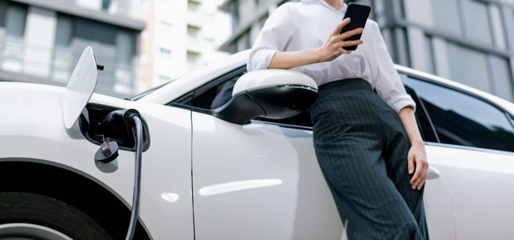 Closeup progressive businesswoman with smartphone, leaning electric car and charging station before driving around city center. Eco friendly rechargeable EV car powered by sustainable and clean energy