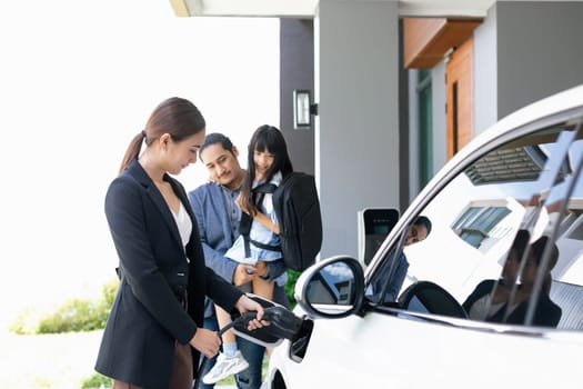 Progressive young parents and daughter with electric vehicle and home charging station. Green and clean energy from electric vehicles for healthy environment. Eco power from renewable source at home.