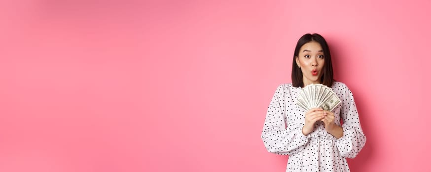 Shopping concept. Excited asian woman holding money, gasping amazed and staring at camera, standing over pink background.