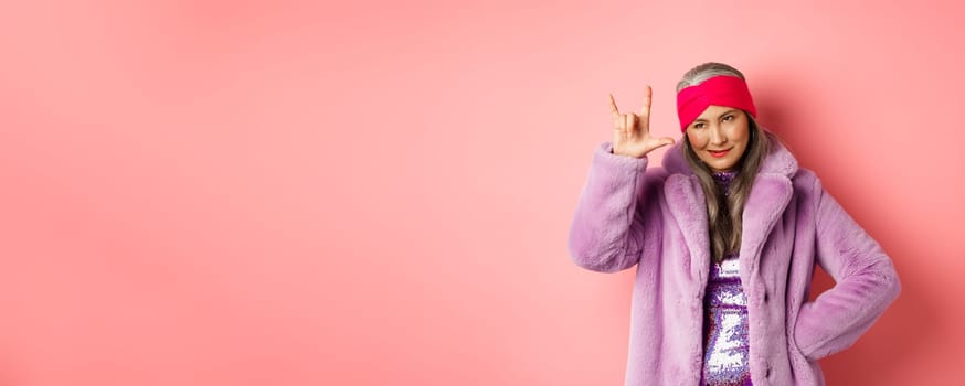 Funny and cool asian senior woman showing rock n roll gesture, looking sassy, standing over pink background.