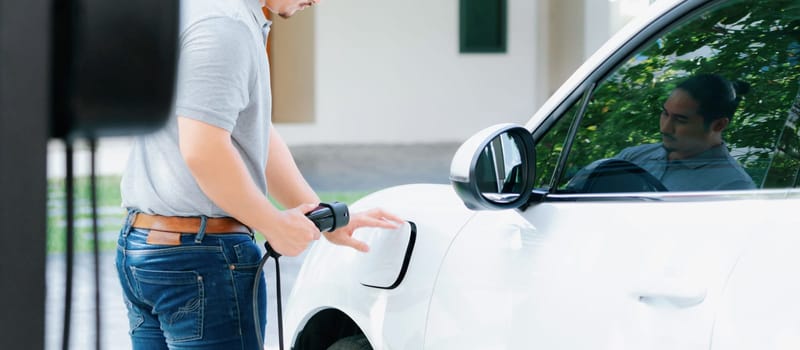 Progressive asian man install cable plug to his electric car with home charging station in the backyard. Concept use of electric vehicles in a progressive lifestyle contributes to clean environment.