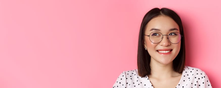 Beauty and lifestyle concept. Close-up of cute asian female model wearing trendy glasses, smiling and looking left at copy space, standing on pink background.