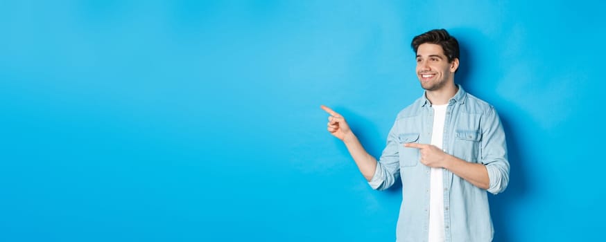 Handsome adult man introduce product, looking and pointing fingers left, promoting something against blue background.