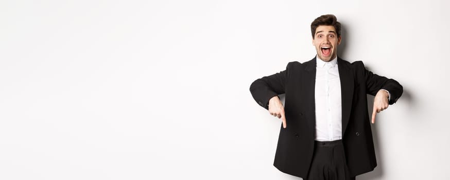 Portrait of super happy man showing christmas promo offer, pointing fingers down and smiling amazed, wearing formal party suit, standing over white background.