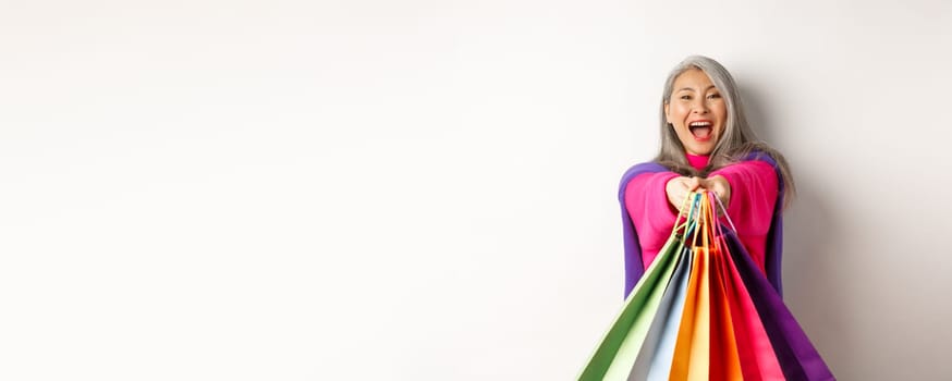 Excited korean lady with grey hair, laughing and showing shopping bags, visit stores with special discounts, standing over white background.