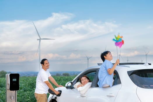 Concept of progressive happy family enjoying their time at wind farm with electric vehicle. Electric vehicle driven by clean renewable energy from wind turbine generator for charging station.