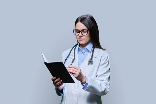 Young serious female doctor writes in notebook, on gray studio background. Confident female in white coat with glasses looking at notebook. Medical staff occupation health care science medicine