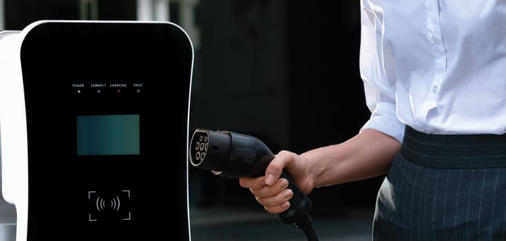 Focus EV charger plug and electric car at public charging station with blur progressive businesswoman holding charger and apartment condo building in background. Eco friendly electric vehicle concept.