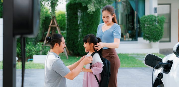 Progressive young parents and daughter with electric vehicle and home charging station. Green and clean energy from electric vehicles for healthy environment. Eco power from renewable source at home.