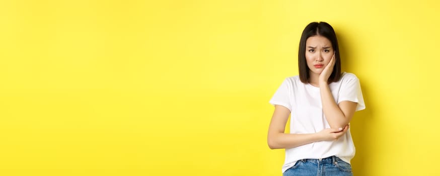 Sad asian woman touching cheek and frowning, having toothache, need see dentist, standing gloomy against yellow background.