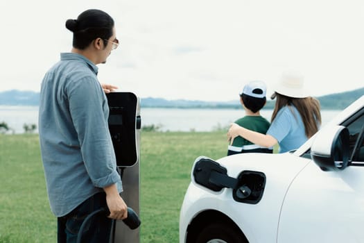 Concept of progressive happy family enjoying their time at green field and lake with electric vehicle. Electric vehicle driven by clean renewable from eco-friendly power sauce.