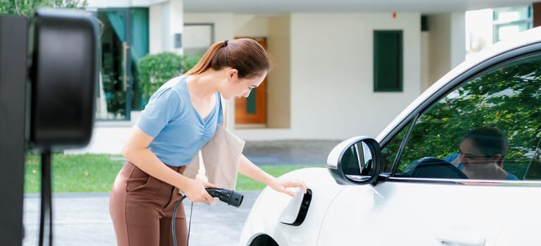 Progressive woman install cable plug to her electric car with home charging station. Concept of the use of electric vehicles in a progressive lifestyle contributes to clean environment.