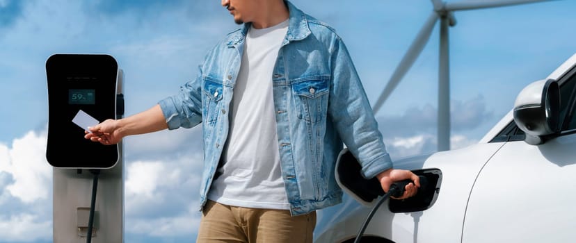 Progressive man with his electric car, EV car recharging energy from charging station on green field with wind turbine as concept of future sustainable energy. Electric vehicle with energy generator.