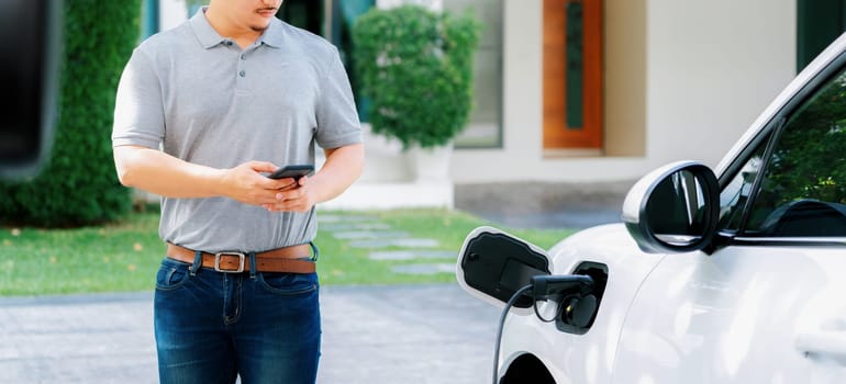 Progressive asian man install cable plug to his electric car with home charging station in the backyard. Concept use of electric vehicles in a progressive lifestyle contributes to clean environment.