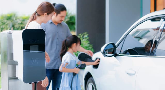 Closeup focus hand insert EV charger plug to electric vehicle at home charging station with blurred family in background. Progressive concept of green and clean energy to reduce CO2 emission by EV car