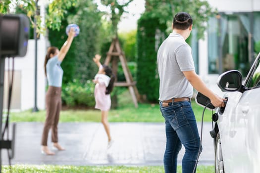 Focus image of progressive man charging electric car from home charging station with blur mother and daughter playing together in the background.