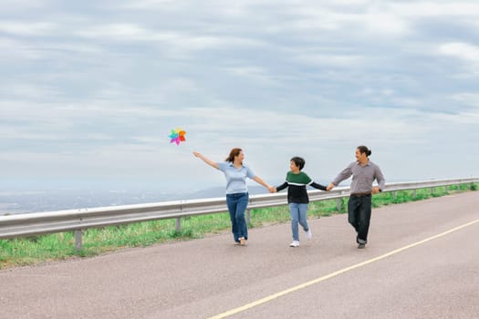 Progressive happy family vacation and carefree day concept. Young parents mother father and son run along and flying kite together road with enjoy natural scenic on scenery and clear sky background.