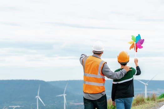 Engineer with his son holding windmill toy on a wind farm atop a hill or mountain. Progressive ideal for the future production of renewable, sustainable energy. Energy generated from wind turbine.