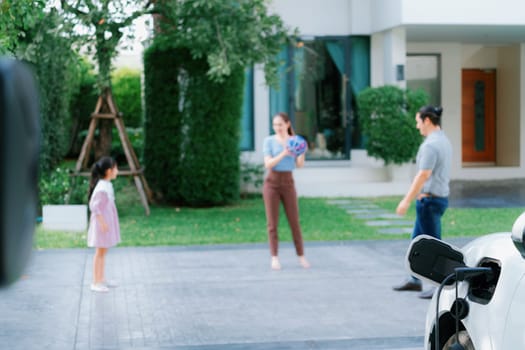 Focus closeup electric vehicle recharging battery from home electric charging station with blurred family in background. Renewable clean energy car for progressive eco awareness lifestyle concept.
