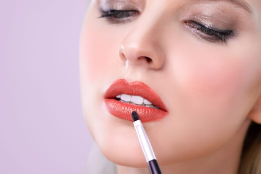 Closeup beautiful young woman with flawless healthy skin and natural makeup putting alluring fashion glossy red lipstick on her lip with lip brush in pink isolated background.