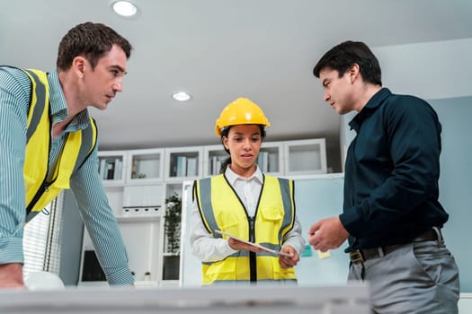 A team of competent engineers wearing safety equipment is working on blueprints with a tablet while also discussing with the investor in the office.