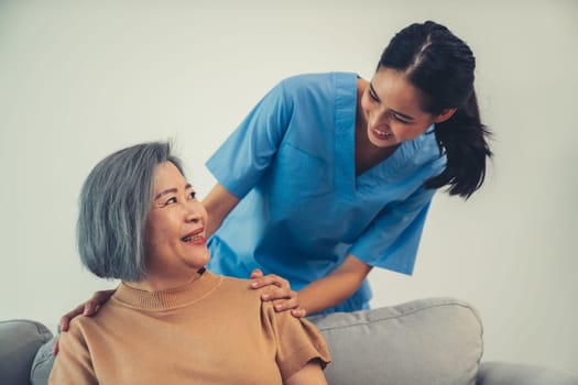 A caregiver rest her hands on the shoulders of a contented senior patient while she sitting on the sofa at home.