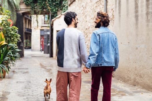 rear view of a gay male couple smiling happy walking with their little dog along a beautiful old street with plants, concept of lifestyle and love between people of the same sex